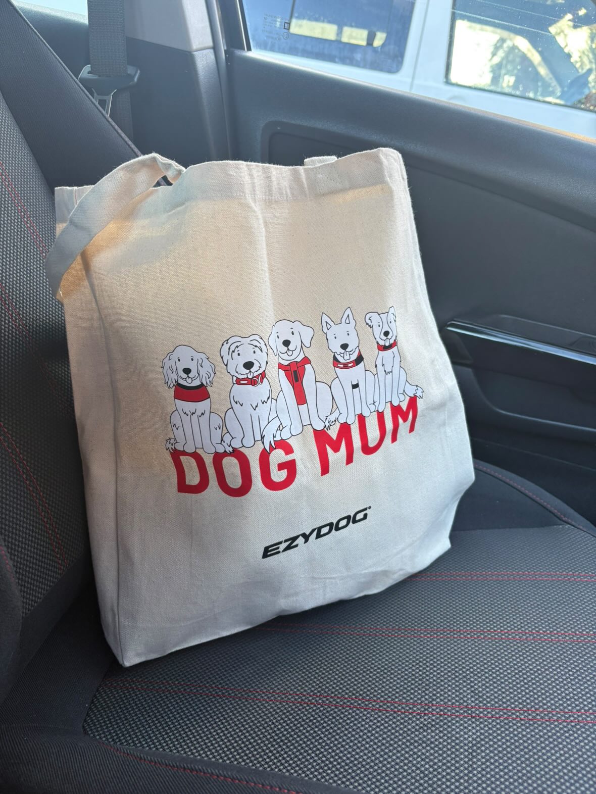 Beige tote bag with 'DOG MUM' and dog illustrations on a car seat.